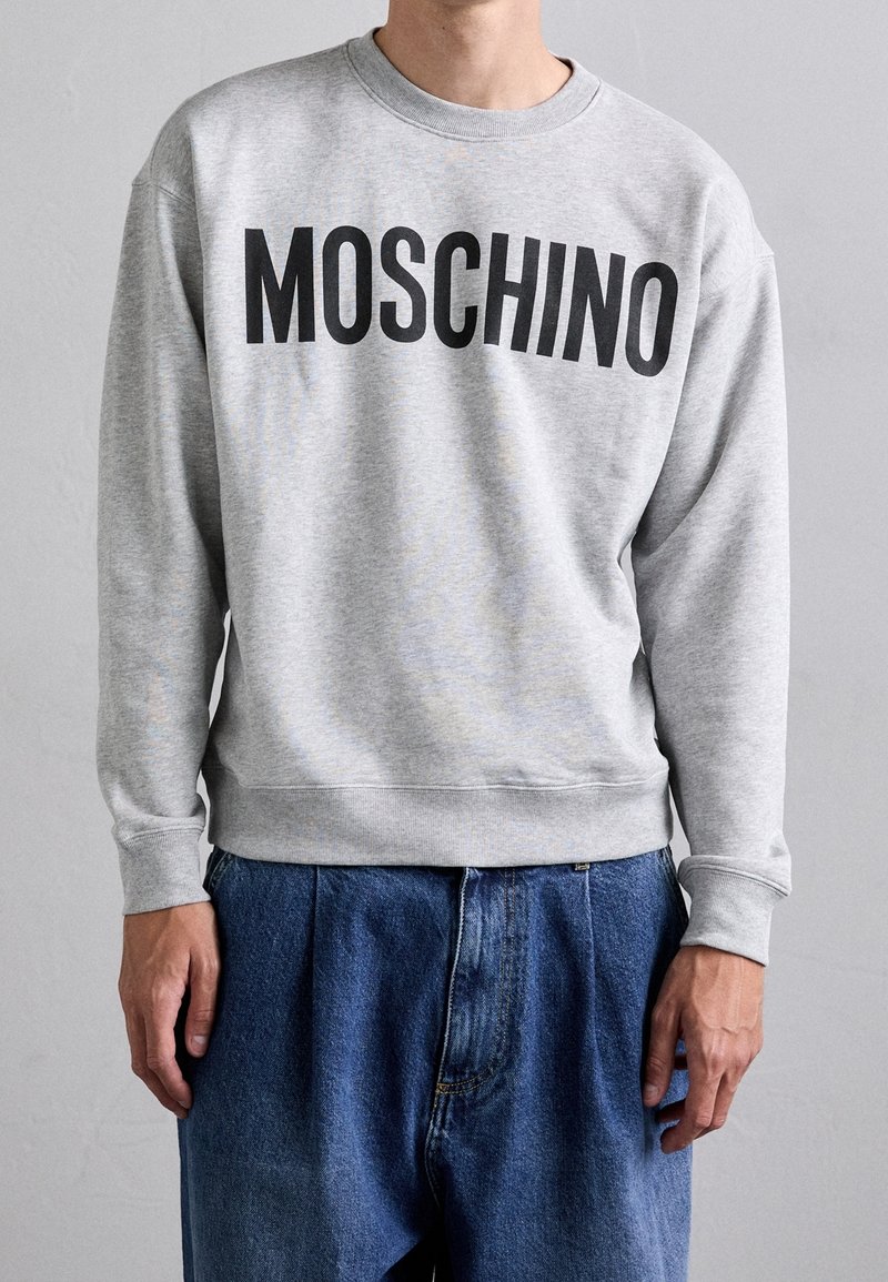 Personne portant un sweat-shirt gris clair avec « MOSCHINO » en grandes lettres noires sur la poitrine et un jean bleu ample.