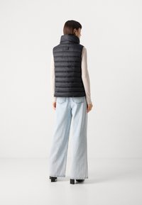 Doudoune noire sans manches avec col haut, design matelassé et détail logo dans le dos, associée à un jean bleu clair à jambes larges. Fond uni.