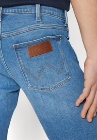 Lichtblauwe spijkerbroek van denim met een leren Wrangler-patch op de achterzak, voorzien van een gestikte "W"-ontwerp en metalen klinknagelaccenten.