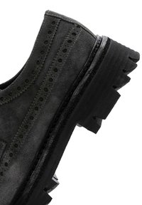 Chaussure en cuir suédé noir avec des détails perforés, semelle en caoutchouc épaisse et bords rugueux. Présente une couture subtile le long du dessus et des accents surélevés.