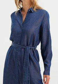 Sukienka koszulowa z denimu w ciemnym niebieskim kolorze, z zapięciem na guziki z przodu, długimi rękawami i paskiem wiązanym w talii. Miękka w dotyku z delikatnym połyskiem.