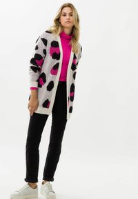 Gebreide cardigan met een grijze basis, zwarte en roze luipaardpatronen, open voorkant, lange mouwen, gecombineerd met een roze coltrui en zwarte broek.