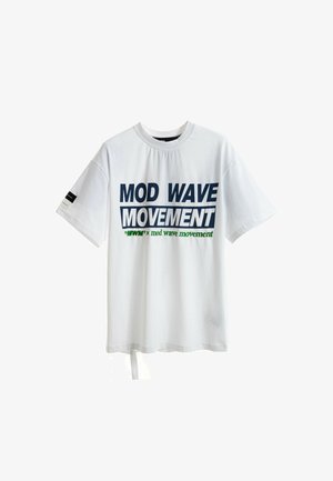 Joven de pie vistiendo una camiseta blanca con la inscripción "MOD WAVE MOVEMENT", pantalones cortos de mezclilla azul, calcetines blancos con estampado y zapatos beige texturizados.