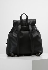 Sac à dos en cuir noir avec un rabat rabattable, deux bretelles et un détail de fermoir contrastant en métal doré. Texture lisse et forme structurée.