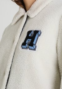 Crèmekleurige faux sherpa jas met een kraag, voorzien van een blauwe en zwarte geborduurde letter "H" accent aan de linkerkant van de borst.