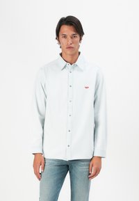Mann i lys blå denimskjorte med rødt logo på brystet og lyseblå jeans, som står mot hvit bakgrunn.