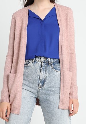 Cardigan a maglia rosa chiaro con apertura frontale, polsini a coste e due tasche frontali, indossato sopra una blusa blu e jeans in denim a vita alta.
