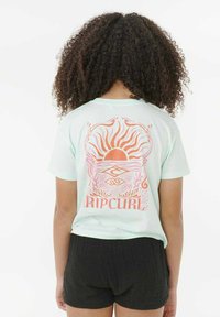 Persona con cabello rizado que lleva una camiseta azul claro de Rip Curl con un diseño de sol naranja y océano en la parte posterior y pantalones cortos negros.