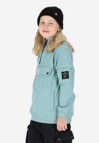 Hellblauer Pullover aus weichem Material, mit einem Reißverschlusskragen, einer Fronttasche und einem schwarzen Logo-Patch am Ärmel. Eine schwarze Mütze wird getragen.