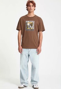 Bruin katoenen t-shirt met een grafisch ontwerp in zwart, wit en goud, gecombineerd met lichtblauwe spijkerbroek en zwart-witte sneakers.