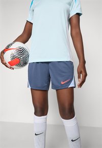 Light blue sports top with short sleeves, paired with blue shorts featuring an orange Nike logo. Player holds a patterned soccer ball.