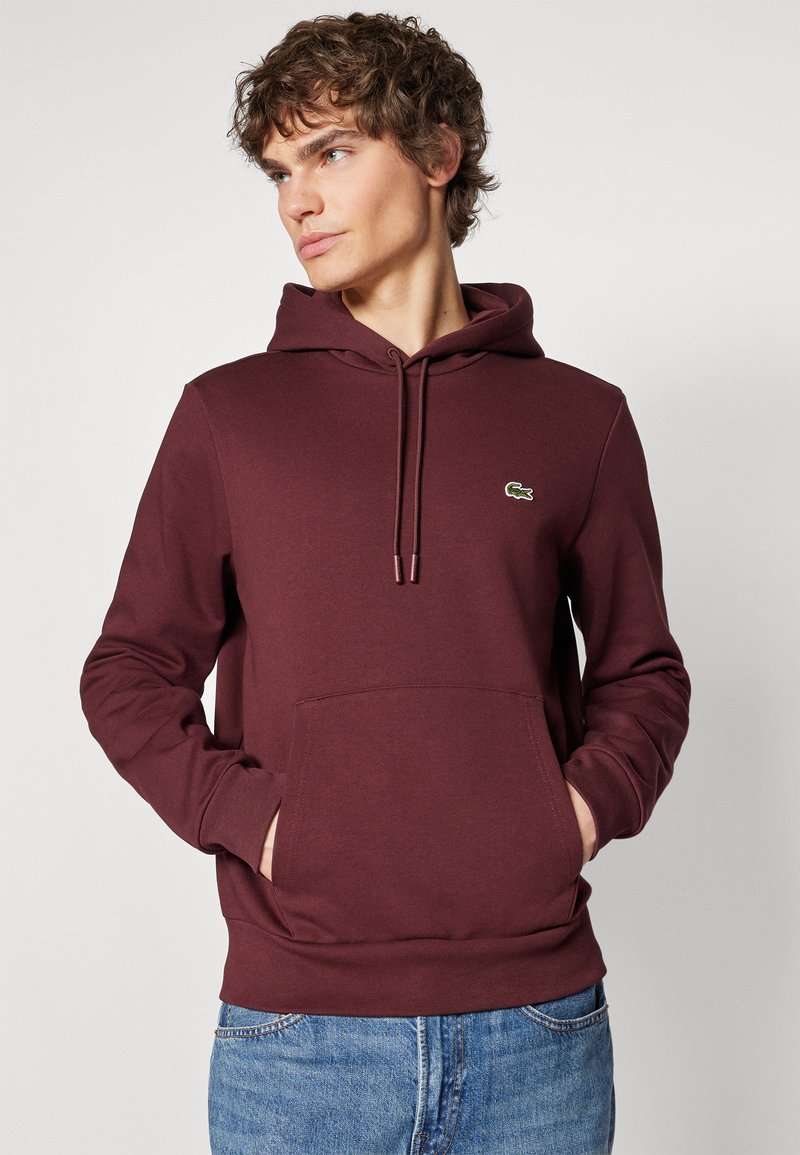 Bordeauxfarbener Hoodie mit Kängurutasche, Kapuze mit Kordelzug und kleinem grünen Logo-Detail. Bequemes Baumwollmischgewebe und gerippte Bündchen.