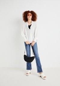 Camicia bianca a maniche lunghe con bottoni, canotta nera sotto, jeans blu a zampa d'elefante, zoccoli bianchi aperti davanti e borsa nera intrecciata. Occhiali da sole rosa.