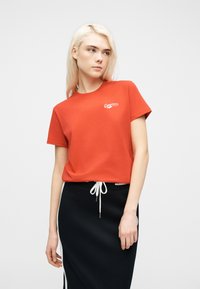 Νέα γυναίκα με ξανθά μαλλιά, ντυμένη με κόκκινο t-shirt Converse και μαύρη φούστα με λευκό κορδόνι, που στέκεται μπροστά σε απλό λευκό φόντο.