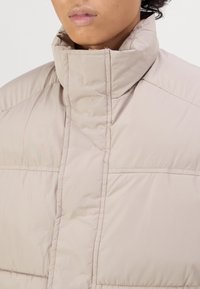 Beige pufferjack met een hoge kraag, voorzien van een gewatteerd ontwerp en gestikte panelen, gladde textuur, geen zichtbare hardware of accenten.