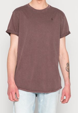 Mann trägt ein verblasstes braunes T-Shirt mit kurzen, hochgekrempelten Ärmeln und hellblauen Jeans und zeigt ein Unterarmtattoo.