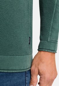 Zelená strukturovaná mikina s designem vaflové pleteniny, žebrovanými manžetami a logem na boční straně poblíž lemu.