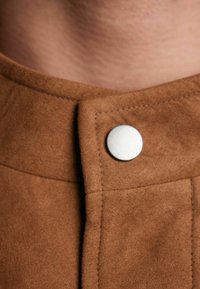 Brązowy zamsz w zbliżeniu z okrągłym srebrnym guzikiem, wysokim kołnierzem i widocznym stębnowaniem na tkaninie. Miękka tekstura i subtelny połysk.