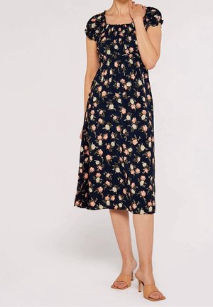 Frau trägt ein dunkelblaues, blumiges Midikleid mit Puffärmeln und quadratischem Ausschnitt, kombiniert mit nudefarbenen, hochhackigen Sandalen.