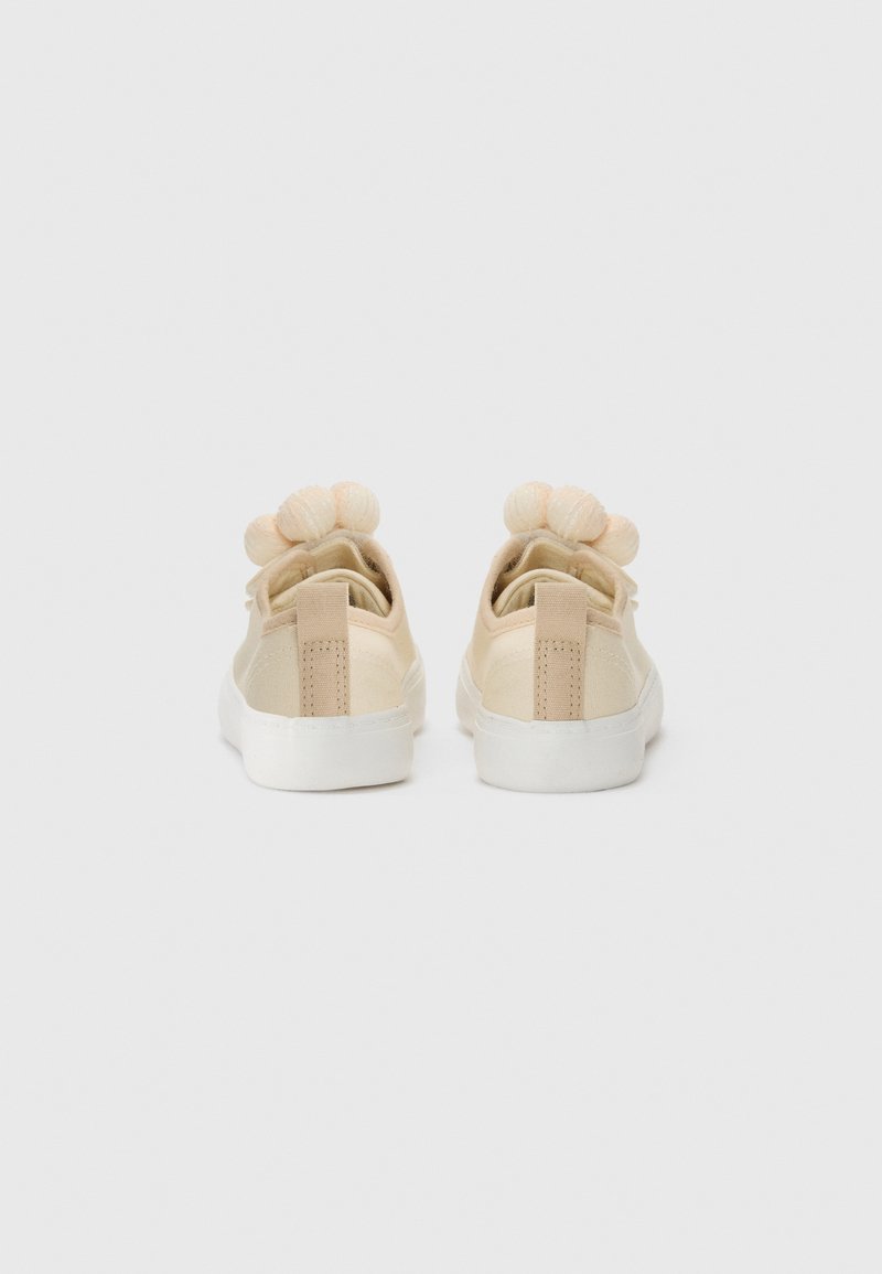 Beige Sneaker aus Canvas mit einer weißen Gummisohle, ausgestattet mit einem strukturierten Rückenteil und drei weichen Pompons oben auf der Ferse.