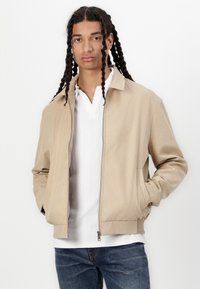 Veste bomber beige avec un col, fermeture éclair sur le devant et poches latérales. Portée par-dessus une chemise blanche texturée et associée à un jean bleu.