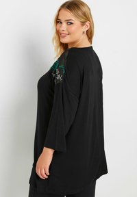 Schwarze Tunika mit 3/4-Ärmeln, verziert mit floraler Stickerei auf transparenten Schulterpaneelen. Weicher Stoff, lockere Passform und abgerundeter Saum.