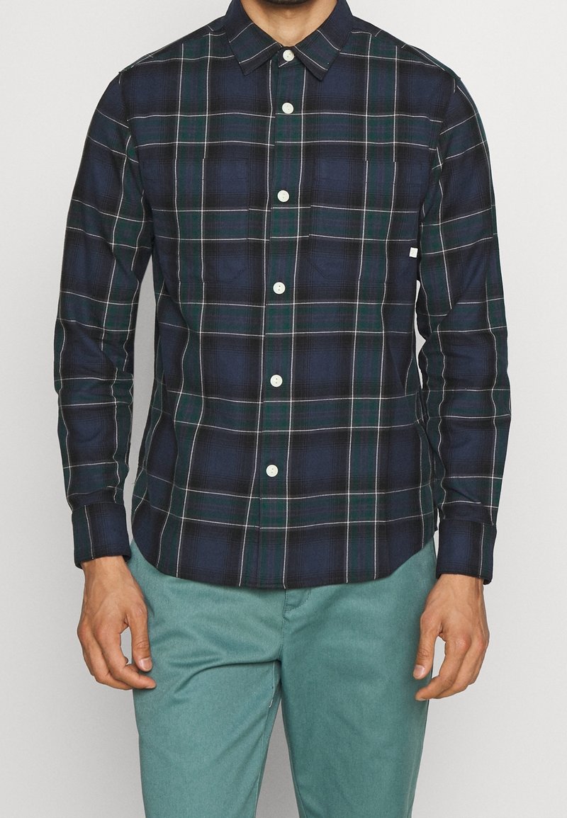 Hombre vestido con una camisa de cuadros en azul marino y verde de manga larga con botones blancos y pantalones en tono teal, posando contra un fondo liso.