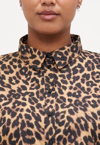 Blouse en imprimé léopard avec col, présentant un fond beige et des taches noires/brunes, et des boutons noirs sur le devant. Texture de tissu lisse.