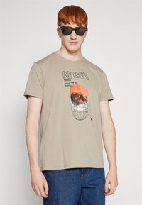 Beige katoenen t-shirt met een NASA graphic design met een oranje zonsondergang en wereldbol. Gecombineerd met een donkere zonnebril en blauwe jeans.