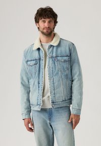 Lichtblauwe denimjack met een met fleece gevoerde kraag, knoopsluiting, twee borstzakken en een losse fit. Draagt over een grijs shirt.