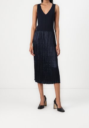 Femme portant une robe noire sans manches à col en V avec une jupe plissée et des chaussures noires à talons hauts, debout sur un fond blanc.