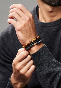 Tre armbånd på et håndledd: ett med trege perler, ett med svarte lavaklipper, og ett med en liten metallisk aksent, alle i ulike farger.