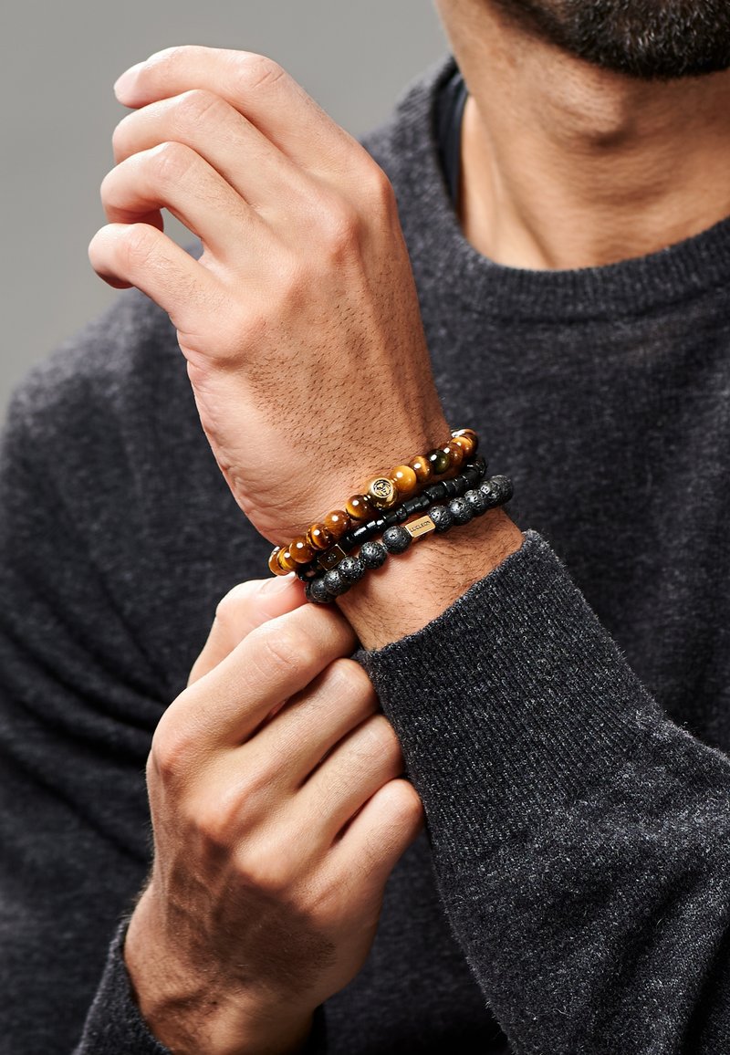 Tre armbånd på et håndledd: ett med trege perler, ett med svarte lavaklipper, og ett med en liten metallisk aksent, alle i ulike farger.