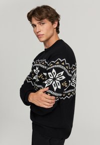 Czarny sweter dzianinowy z białymi i złotymi wzorami płatków śniegu na rękawach i klatce piersiowej. Okrągły dekolt i ściągacze przy mankietach.