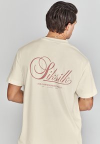 Camiseta de algodón color crema con un gran diseño burdeos en la parte posterior que incluye el texto "Siksilk" y "FOLLOWTHEMOVEMENT" debajo.