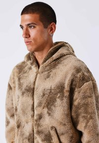 Giovane uomo con capelli corti che indossa una giacca in pile a zip beige e marrone con cappuccio e un piccolo logo sul petto.