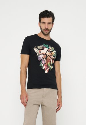 Bărbat purtând un tricou negru cu un design geometric de urs înconjurat de crenguțe de pin, asortat cu pantaloni bej, stând pe un fundal alb.