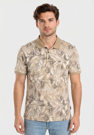 Giovane uomo con polo beige a motivo foglia e jeans blu, in piedi davanti a uno sfondo chiaro e uniforme, rivolto verso l'osservatore.