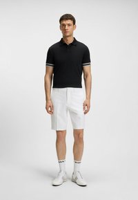 Polo nero con maniche a righe bianche, abbinato a pantaloni corti bianchi. Il modello indossa sneaker bianche e calze bianco e nere, in piedi su uno sfondo chiaro.