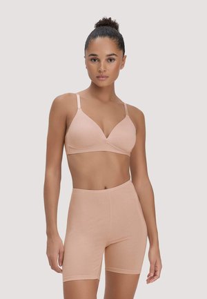 Femme aux cheveux bouclés attachés en chignon, portant un soutien-gorge couleur nude et un short cycliste taille haute, sur un fond uni et clair.