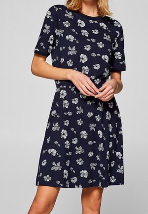 Nainen, joka käyttää tummansinistä lyhythihaista mekkoa, jossa on valkoisia kukkakuvioita, seisoo kädet löyhästi vyötäröllä.