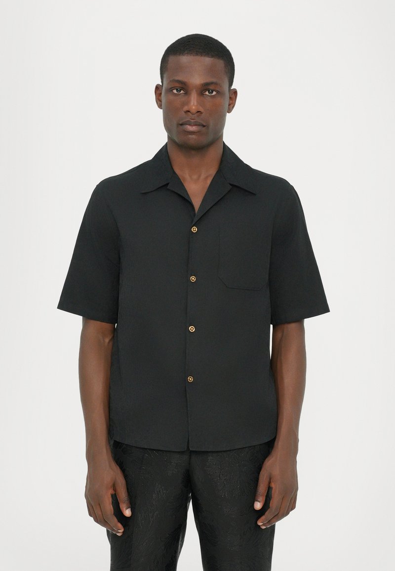 Chemise noire à manches courtes avec un col, poche avant et sept boutons dorés. Fabriquée dans un tissu lisse, avec un design simple.