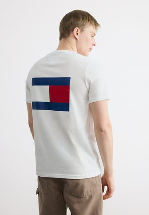 Νέος άνδρας με λευκό μπλουζάκι με μεγάλο κόκκινο, λευκό και μπλε ορθογώνιο σχέδιο στην πλάτη, φοράει καφέ παντελόνι, στέκεται πλευρικά.