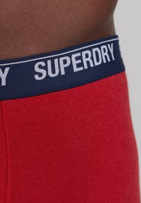 Rode boxershorts met een marineblauwe elastische tailleband waarop het woord "SUPERDRY" in het wit staat. Gladde textuur, close-up weergave.