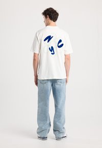 White short-sleeve t-shirt with blue and green "NYC" graphic design on the back. Worn with light blue denim jeans.