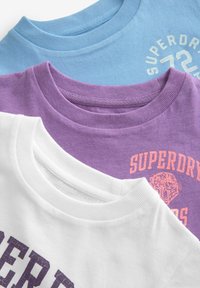 Tre crewneck T-shirts i blå, lilla og hvid. Den lilla T-shirt har kontrastfuld pink tekst og et tigermotiv; de andre er ensfarvede.