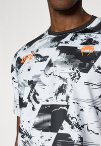 Αθλητική μπλούζα με κοντά μανίκια σε λευκό και μαύρο χρώμα, με γεωμετρικό αφηρημένο σχέδιο και πορτοκαλί λογότυπο "UFC" στο μπροστινό μέρος. Λείο υλι κό.