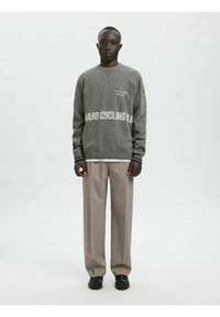 Grauer Wollpullover mit den Schriftzügen "LA FLAMME ROUGE" und "HARD CYCLING", getragen über einem weißen Hemd, kombiniert mit beigen Hosen und schwarzen Schuhen.