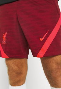 Viininpunaiset urheilushortsit, joissa on teksturoitu kuvio, Liverpool FC -logo sekä punainen Nike-viiva sivussa. Henkilön alaleuka on näkyvissä.
