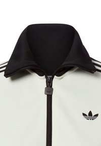 Chaqueta blanca con cremallera y cuello negro, con tres rayas negras en cada hombro y un pequeño logo negro en el pecho.
